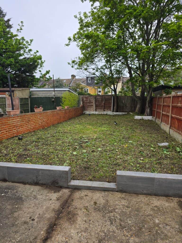 A rectangular backyard with patchy grass, bordered by red wooden fences and a low brick wall, with trees and some houses visible in the background.