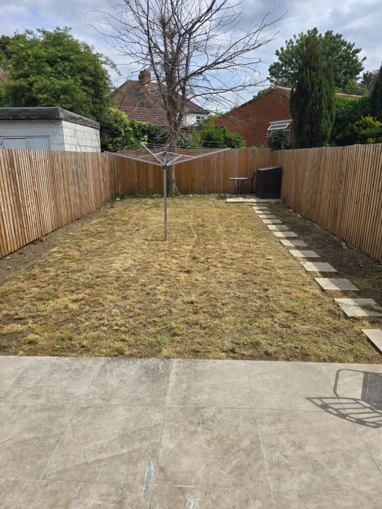 A fenced backyard with a patchy lawn, a leafless tree in the center, stone path on the right, and outdoor furniture near the fence in the background.