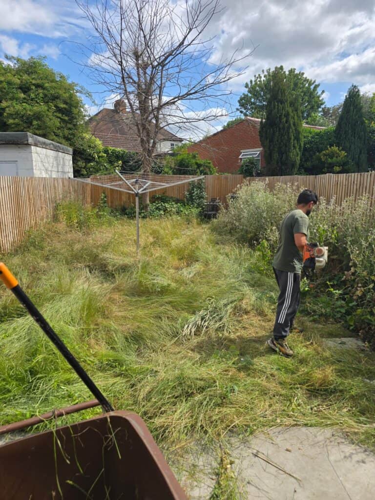 A person in a green shirt and black pants is using a trimmer to cut overgrown grass and weeds in a backyard with a brown bin and rotary clothesline visible.