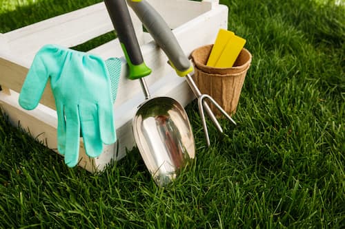 A set of gardening tools, gloves, and a small pot are placed on grass next to a white wooden crate.